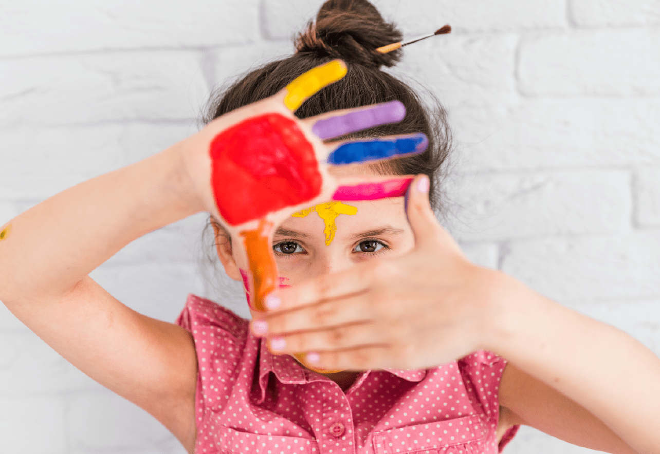 Niña mirando a cámara ocultando parte del rostro con sus manos manchadas de pintura