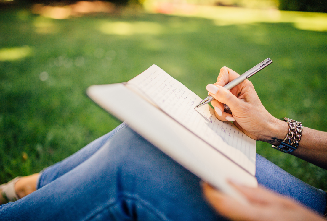 Unas manos de mujer escribiendo en una libreta que tiene sobre las piernas