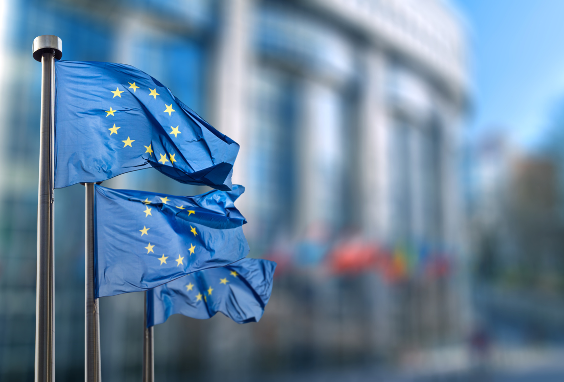 Primer plano de unas banderas europeas con el edificio del Parlamento Europeo de fondo